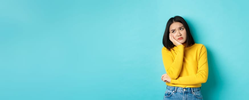 Sad and bored asian teenage girl leaning on palm, looking at upper left corner with lonely and upset expression, standing over blue background