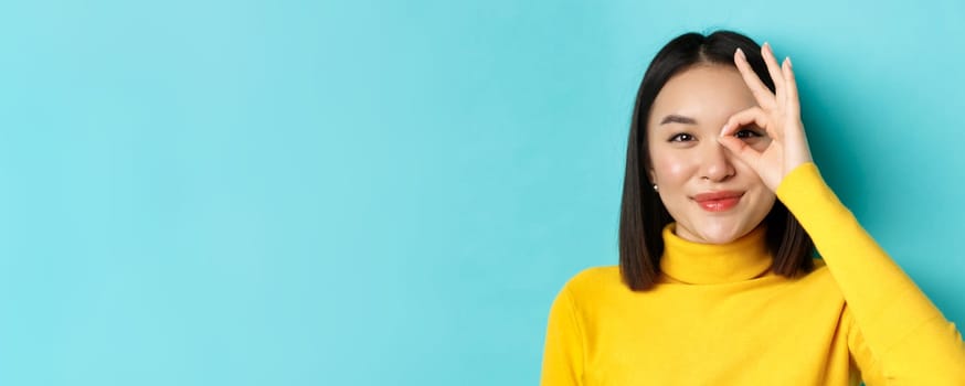 Beauty and makeup concept. Close up of carefree asian girl showing OK sign on eye and smiling, looking happy at camera, standing over blue background