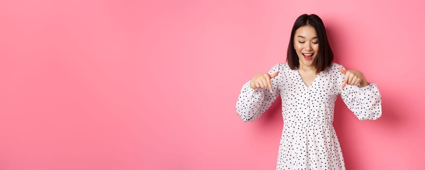 Romantic asian woman peeking down, pointing fingers at bottom, looking curious at product discount with happy smile, standing over pink background