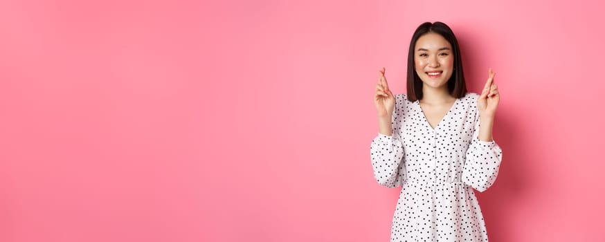 Cute asian woman making a wish, smiling happy and cross fingers for good luck, waiting over pink background