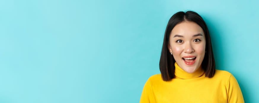 Beauty and skincare concept. Close up of surprised asian female raising eyebrows and look amused at camera, look at something interesting, standing over blue background