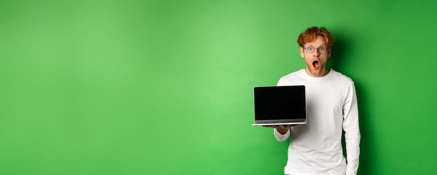 Impressed redhead guy in glasses drop jaw after seeing online promo, showing laptop screen and staring at camera with disbelief, standing over green background