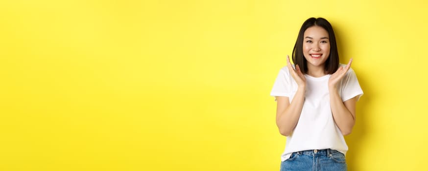 Beauty and fashion concept. Excited asian woman clap hands and smiling happy at camera, standing in white t-shirt against yellow background