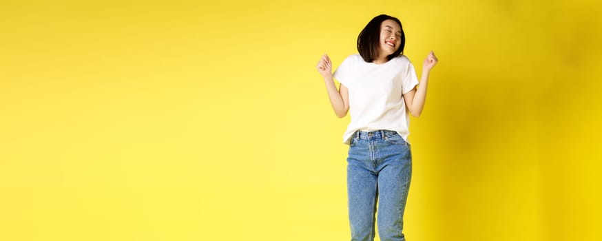Full size shot of carefree asian woman jumping and dancing, having fun, posing in jeans and white t-shirt over yellow background