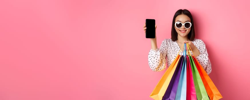 Attractive asian woman showing smartphone app and shopping bags, buying online via application, standing over pink background