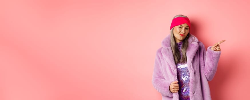 Displeased mature asian woman in stylish purple fake-fur coat pointing finger right, grimacing from dislike, looking skeptical and disappointed, pink background