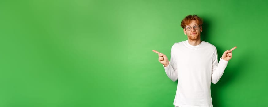 Indecisive funny guy with red hair pointing fingers sideways, staring confused at camera and showing two promo offers, standing over green background