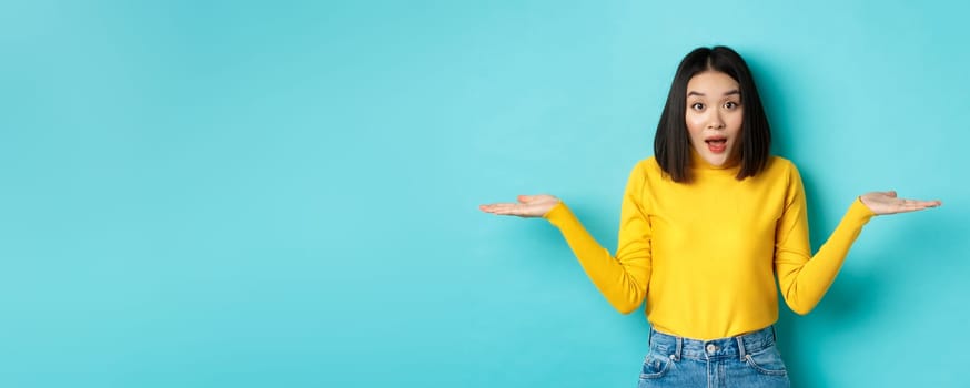 Image of indecisive asian woman shrugging shoulders, spread hands sideways and looking clueless at camera, standing confused against blue background