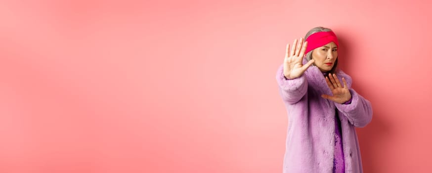 Asian senior female model in stylish purple winter coat standing in victim pose, extending hand in stop gesture and pleading for mercy, standing against pink background