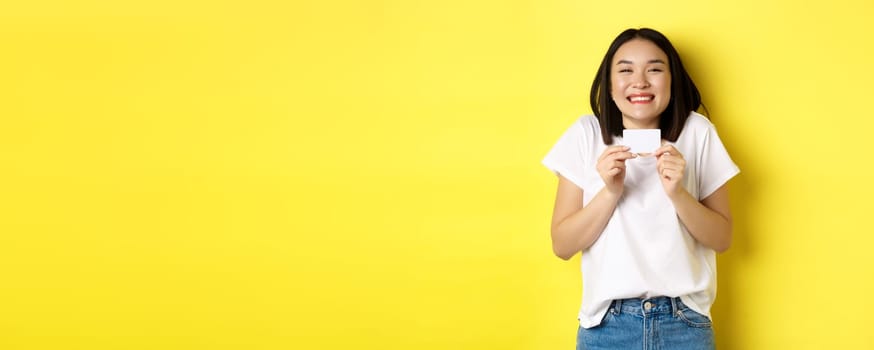 Young asian woman in casual white t-shirt going on shopping, showing plastic credit card and smiling excited, standing over yellow background