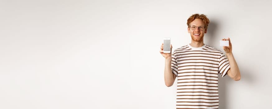Shopping and finance concept. Young man showing blank mobile screen and plastic credit card, smiling at camera, white background