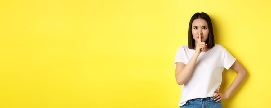Beauty and fashion concept. Angry and bossy asian girl telling to be quiet, scolding loud people, shushing and frowning at camera, standing in t-shirt over yellow background