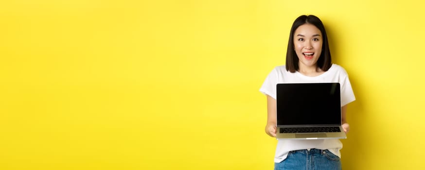 Young asian woman demonstrate online offer, showing blank laptop screen and smiling, standing over yellow background