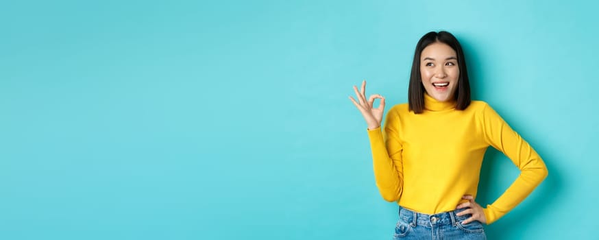 Impressed asian woman in yellow sweater, looking left amazed and showing OK sign in approval, standing over blue background