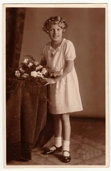 Vintage photo shows young girl with bouquet poses in a photography studio. Photo with dark sepia tint. Black white studio portrait.