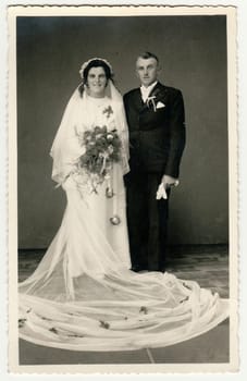 Vintage photo of newlyweds. Bride wears veil, long wedding gown and holds wedding bouquet. Groom wears black suit and white bow tie. Black white antique studio portrait.