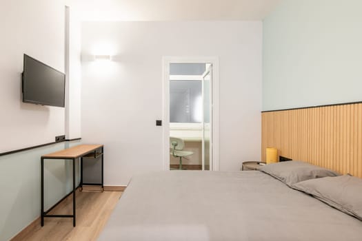 Compact stylish bedroom in stylish soothing colors with a double bed, TV with access to closed balcony with an office area. Concept of a small cozy bedroom of young family.