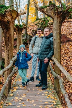 dad and 2 sons in the autumn forest stand on the bridge. Happy family enjoy the spending vacation together in the nature. father and son hug in the wooden pier at the background river and fall leaves forest