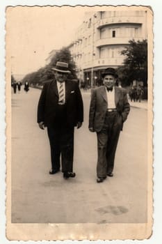 Vintage photo shows the elegant men at the spa resort. They wear hats and slack suits. Colonnade is on background.