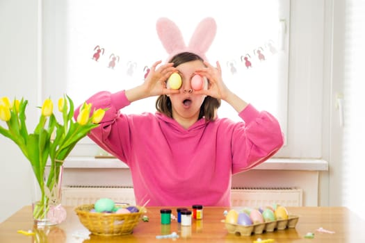Young woman wearing bunny ears having fun while painting Easter eggs at home. Preparation for Easter