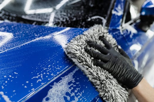 Hand washing with microfiber glove with foam car body in garage. Car washer doing manual foam washing in auto detailing service.