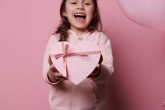 Details on a cute pink heart shaped gift box in the hands of a pretty child girl, smiling over isolated pink background