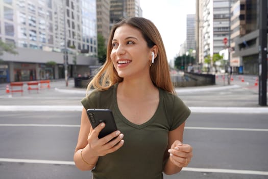 Cute Brazilian Caucasian girl with earphones wireless and mobile phone looks to side in modern metropolis. City life concept.