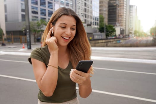 Excited young woman touching her wireless earphones to turn them on and to listening favorite music outdoors
