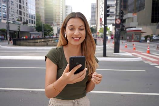 Excited Brazilian girl wearing earphones watching her smartphone in city street