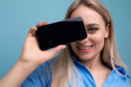 smiling stylish blond girl holding smartphone screen with mockup for web page near her face on blue background