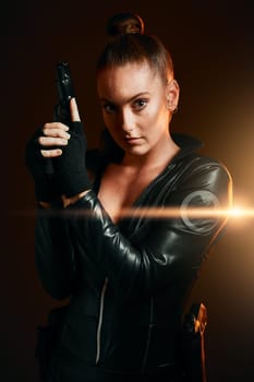 Portrait, gun and secret agent with a woman assassin in studio on a dark background ready for combat. Hero, leather and power with an attractive young female spy holding a weapon on a mission