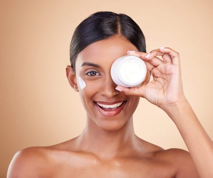Beauty, face cream and portrait of a woman in studio for skincare, dermatology and cosmetics. Happy Indian female model with smile and lotion on hand for self care moisture facial on brown background