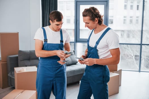 Holding money. Two young movers in blue uniform working indoors in the room