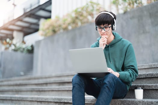 Smart Asian male college student wearing headphones, using laptop on campus outdoor stairs