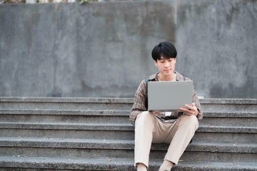 Smart Asian male college student wearing headphones, using laptop on campus outdoor stairs