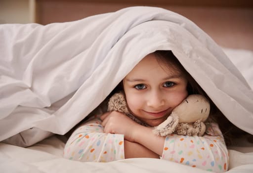 Lovely child girl picking out from soft blanket, cutely smiling looking at camera, gently hugging her cute plush toy