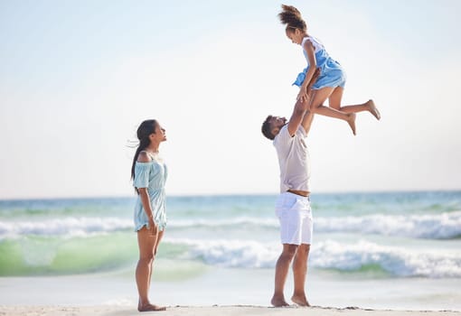 Full length of a happy mixed race family enjoying family time together at the beach. Loving father lifting his daughter in the air and having fun by the sea while her mother watch. Young couple enjoy.