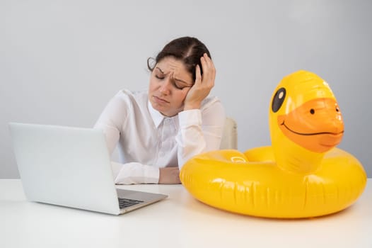 Caucasian woman sits at a table with a laptop and an inflatable duck on a white background. Office worker dreaming of vacation.