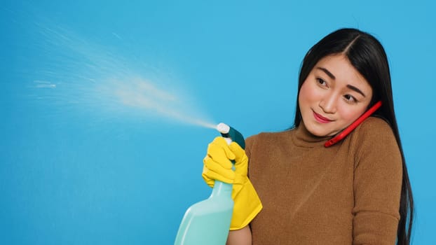 Asian housekeeper using chemical detergent and duster to clean house while talking at mobile phone