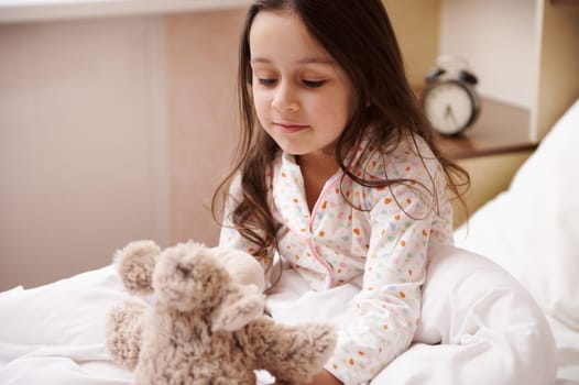 Adorable child girl in pajamas, sitting on bed after waking up in morning, playing with a plush toy in her cozy bedroom