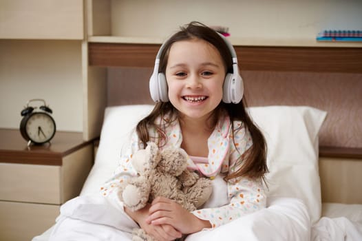 Lovely little girl in pajama and wireless headphones, smiles at camera, sitting on the bed with a plush toy in her hands