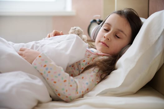 Adorable child girl in headphones, napping in a white bedroom, falling asleep to soothing music on her comfortable bed