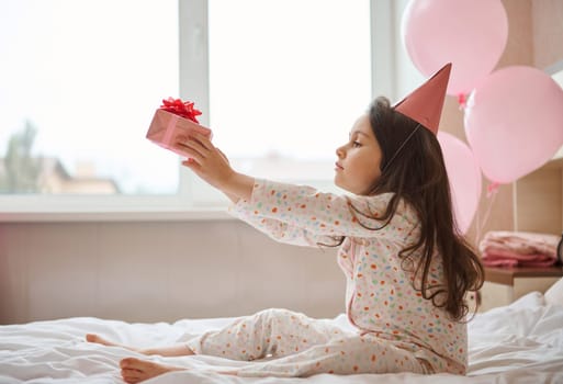 Adorable birthday girl in pink party hat, sitting on the bed in her bedroom and holding her birthday present in hands