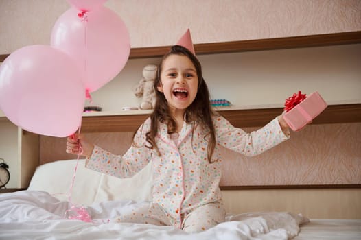 Cheerful birthday girl in party hat, holding bunch of pink balloons and gift box, sitting on bed and smiling into camera