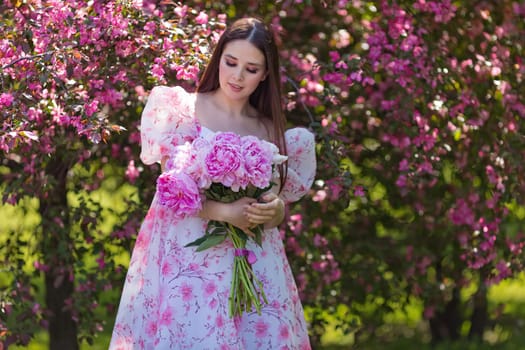 pretty brunette in a light pink dress, with a large bouquet of pink peonies, stands near pink blooming apple trees,in the garden on a sunny day. Copy space.
