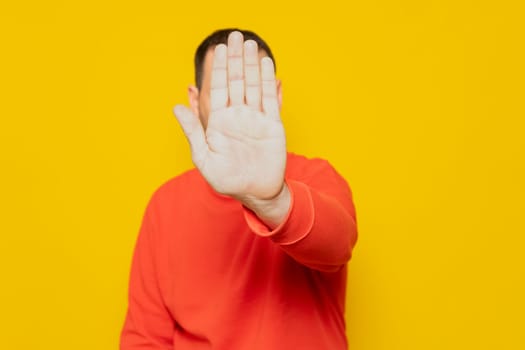 Hispanic man putting hand in front to stop camera, rejecting photos or images against yellow colored wall. Rejection and privacy concept