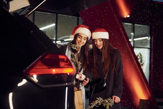 Standing near car's back trunk opened. Happy sisters twins spends Christmas holidays together outdoors. Conception of new year
