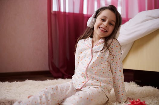 Little child girl in wireless headphones, sitting on a carpet, dressed in stylish pajamas, listening to soothing music