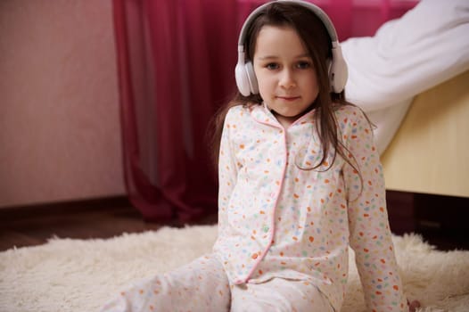 Cute child girl in stylish pajamas, wearing wireless headphones, looking at camera, sitting on the carpet in her bedroom