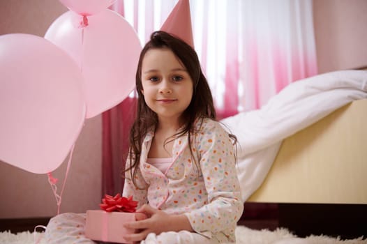 Happy little child girl in pajamas and pink party hat, holds a cute gift box for birthday and smiles looking at camera